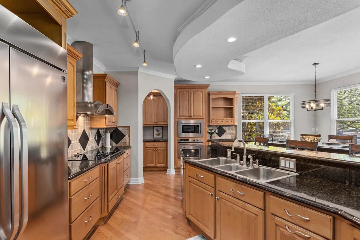 A bright, modern kitchen with a central island, stainless steel appliances, granite countertops, and wood cabinetry, all bathed in natural light from large windows. The space is clearly designed for meal preparation and entertaining groups.