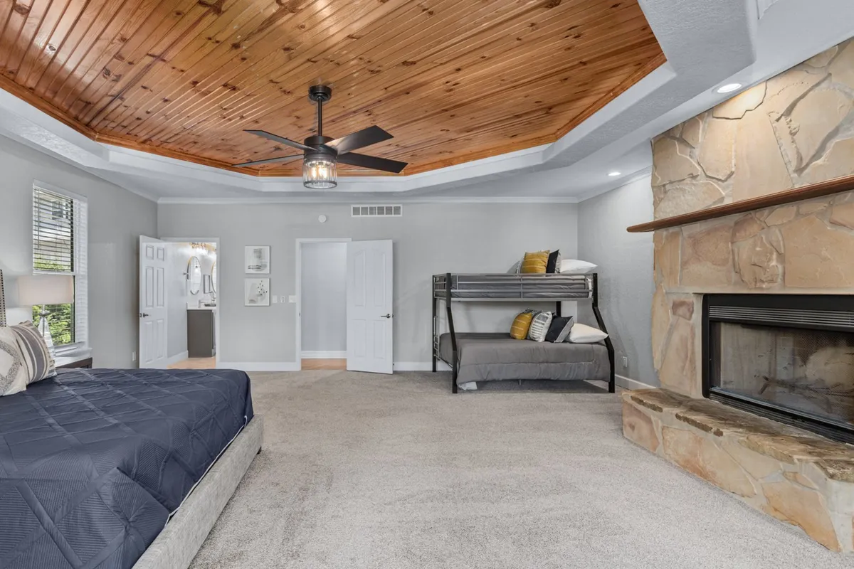 A spacious bedroom featuring a large bed next to a cozy stone fireplace, a wooden ceiling, and a twin/full bunk bed situated along one wall. The room is bathed in soft, natural light from large windows, creating a warm and inviting atmosphere ideal for both families and adults seeking comfort and privacy.