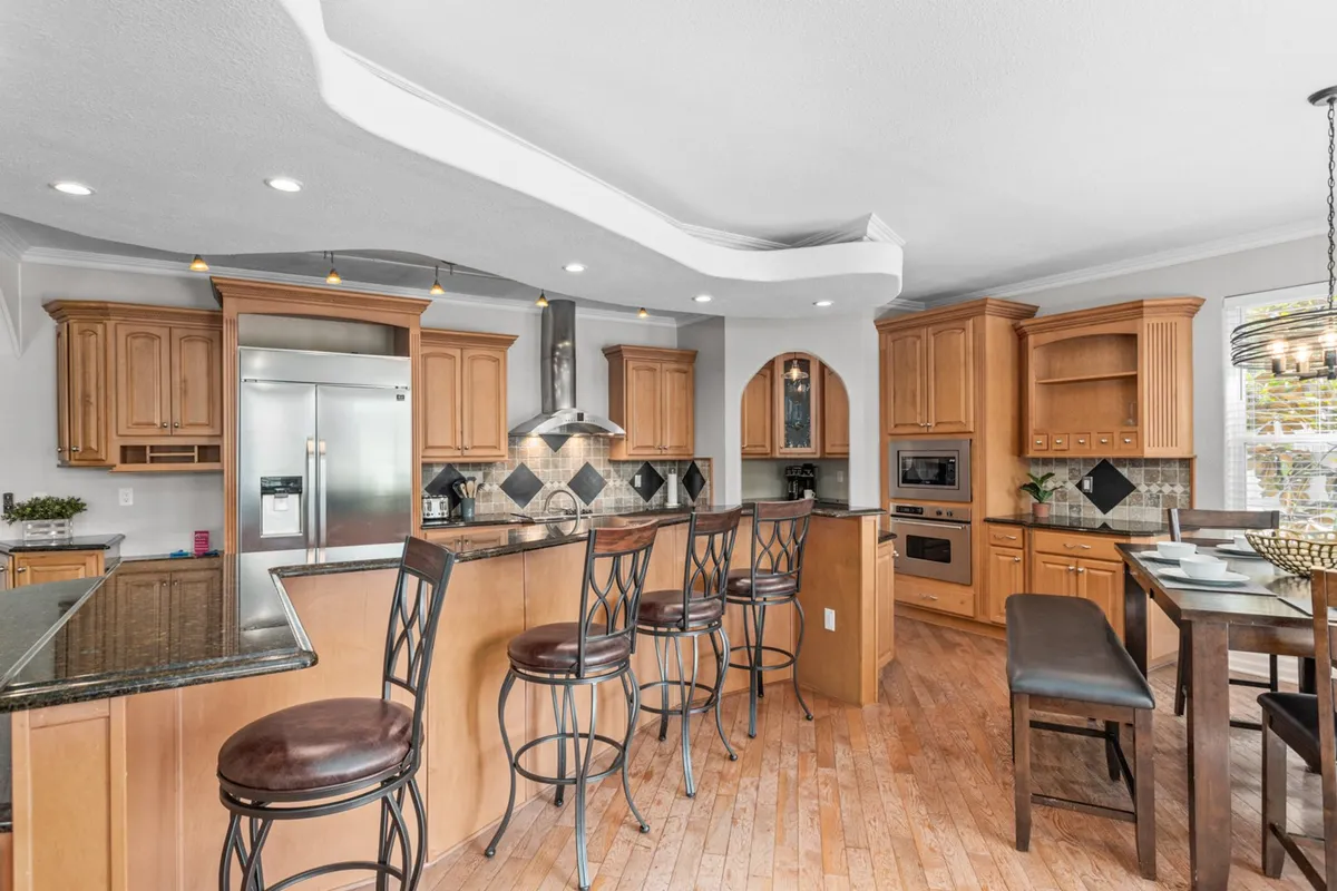 This image features a modern kitchen with a large island surrounded by bar stools, stainless steel appliances, wood cabinetry, and a generous dining table nearby. The layout emphasizes space for both meal preparation and casual dining, with ample natural light creating a warm, inviting feel.