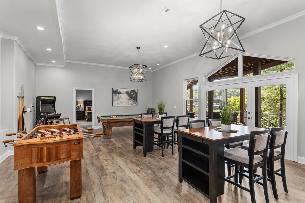 A lively game and family room featuring a foosball table and pool table as central attractions. Bar-style pub seating lines one side, and large windows in the background let in ample natural light, adding warmth to the energetic atmosphere. The room is arranged for social interaction and fun, inviting guests to gather, play, and relax.