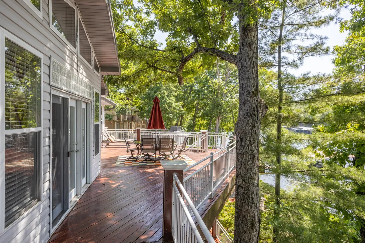 A spacious deck furnished with comfortable seating and a large umbrella, surrounded by lush greenery and overlooking the nearby water. The scene evokes relaxation and social gatherings, with enough space for outdoor dining or enjoying sunset views.