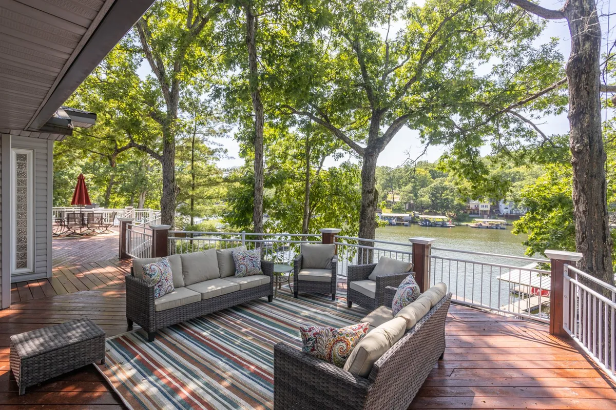 The image showcases a serene outdoor seating area, complete with a decorative rug, nestled under tall trees and overlooking a peaceful cove on the lake. The scene is set for relaxation, with the tranquil water just steps away, inviting guests to unwind, enjoy nature, and bask in beautiful sunset views.