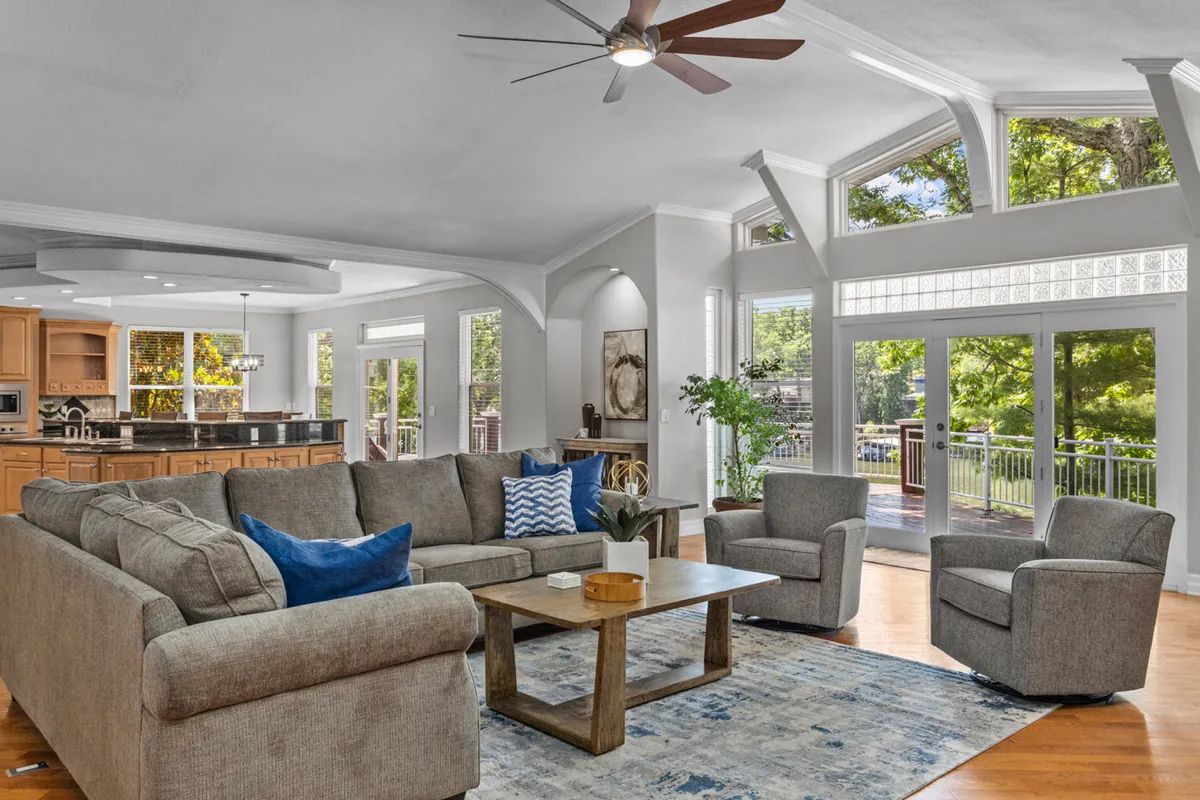 This image showcases a spacious living room with a large sectional sofa and wooden coffee table, oriented around large windows that flood the space with natural light. In the background, the open kitchen area is visible, demonstrating the open-concept layout. The wood floors and plush seating create a cozy, welcoming atmosphere, ideal for gatherings and relaxation.