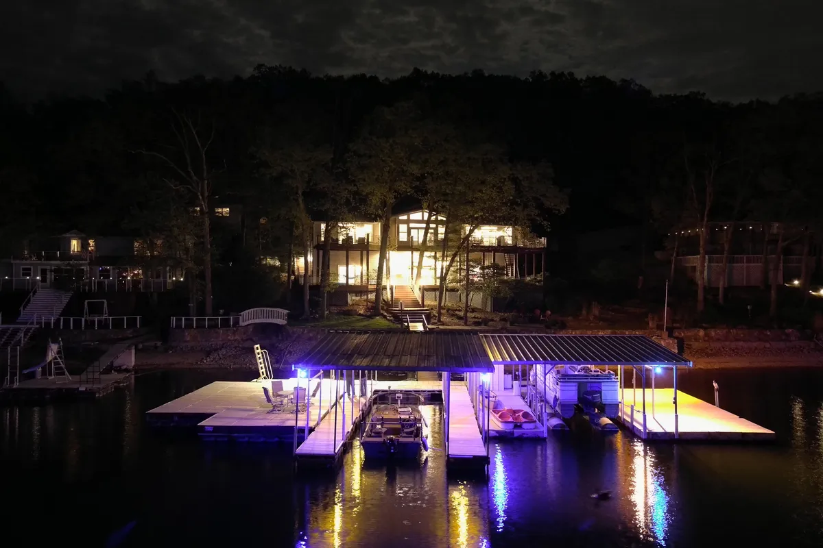 A warmly illuminated house at dusk, its lights reflecting on the still water and a nearby dock. The scene is inviting and cozy, hinting at evenings spent outdoors and the allure of stargazing by the lake.