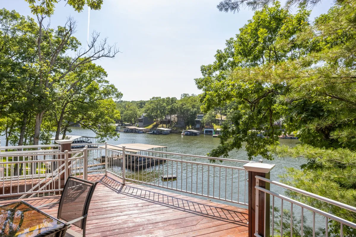 A spacious deck overlooks the calm lake waters, framed by mature trees and dotted with comfortable outdoor furniture. The view includes boats bobbing on the water, evoking a sense of relaxation and easy enjoyment. The setup is perfect for outdoor meals, morning coffee, or watching the sunset, directly tying into the indoor-outdoor lifestyle described.